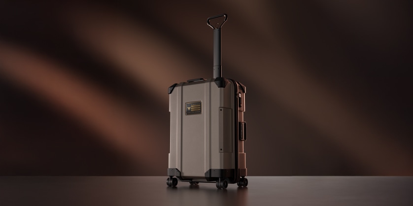 A modern, hard-shell rolling suitcase in a muted gray or beige color is shown standing upright on a dark surface against a blurred brown background.
