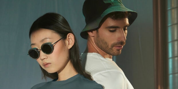Two models, one with dark hair wearing sunglasses and a blue t-shirt, the other with a green bucket hat and white t-shirt with a blurred background.