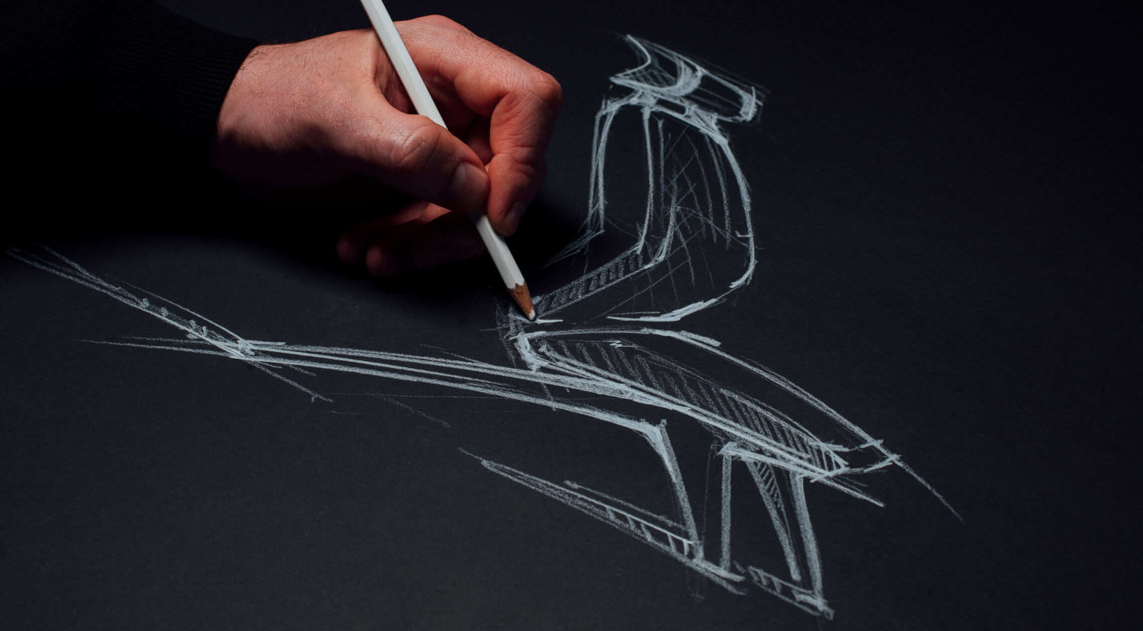 A hand holding a white pencil drawing a sketch of a chair with white lines on black paper.